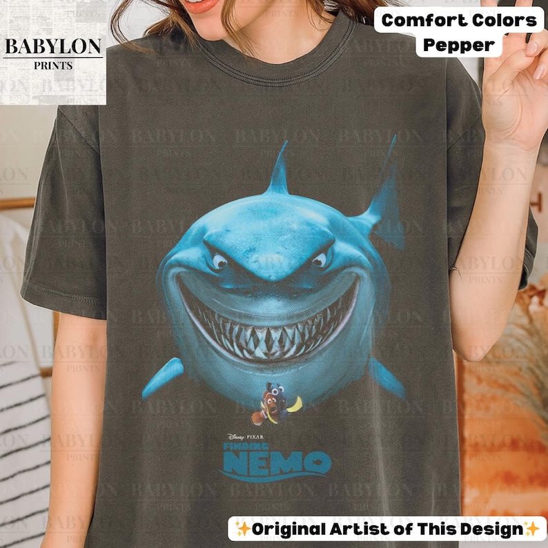 May include: A dark gray t-shirt featuring a large, smiling blue shark graphic from the movie "Finding Nemo." The shirt includes the film's title and the text "Comfort Colors Pepper" and "Original Artist of This Design."