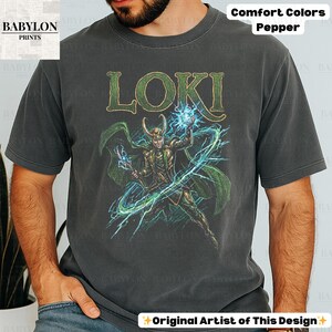 May include: Dark gray t-shirt featuring a graphic of Loki, with the name "LOKI" in green letters. The design includes a character illustration with green and gold accents, and blue lightning effects. The shirt is labeled "Comfort Colors Pepper."