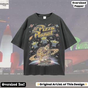 May include: Dark gray oversized t-shirt featuring a colorful graphic of the Pizza Planet logo, aliens, a rocket ship, and planets. The text "Pizza Planet" is in yellow. The shirt also has the text "Oversized Tee!" and "Original Artist of This Design".