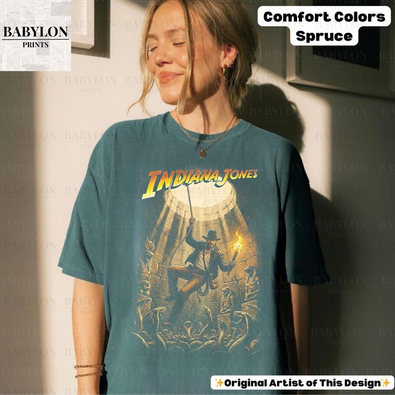 May include: A teal-colored t-shirt featuring the text "INDIANA JONES" in yellow and orange. The graphic depicts a scene from the movie with a figure holding a torch. The shirt also has the text "Comfort Colors Spruce" and "Original Artist of This Design".