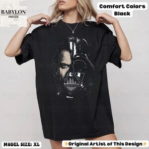 May include: Black t-shirt featuring a split face design of a character from Star Wars. One side shows a human face, the other side shows the character's helmet. The shirt has the text "Comfort Colors Black" and "Original Artist of This Design". The model is wearing size XL.