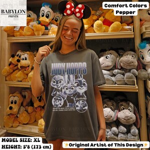 May include: A gray Comfort Colors t-shirt featuring Judy Hopps from Zootopia. The shirt has a graphic of the characters and the text "Judy Hopps" and "Zootopia". The model is wearing a size XL and is 5'8" (173 cm) tall.