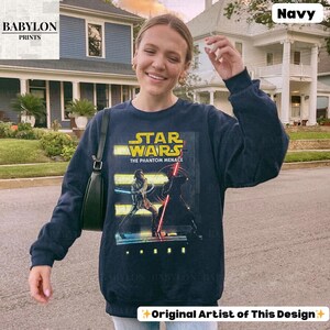Puede incluir: Sudadera azul marino de Star Wars: The Phantom Menace. El gr&aacute;fico presenta el logotipo de Star Wars en amarillo y el t&iacute;tulo de la pel&iacute;cula. La imagen muestra dos personajes con sables de luz. La sudadera es de Babylon Prints.