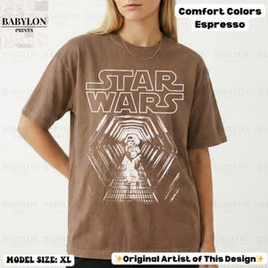 May include: Espresso-colored t-shirt featuring a Star Wars graphic. The design includes the Star Wars logo and a detailed image of a scene from the movie. The shirt is a comfort color and the model is wearing size XL.