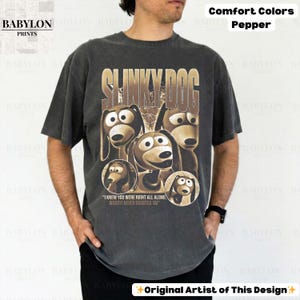 May include: A dark gray t-shirt featuring a graphic of Slinky Dog from Toy Story. The design includes the text "SLINKY DOG" and the quote "I KNEW YOU WERE RIGHT ALL ALONG WOODY! NEVER DOUBTED YA!" The shirt is labeled "Comfort Colors Pepper."