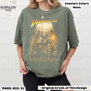 May include: Olive green t-shirt featuring the Indiana Jones movie logo and a graphic of the character. The shirt has short sleeves and a crew neck. The text "Comfort Colors Moss" is printed at the top.