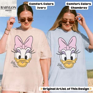 May include: Two t-shirts, one ivory and one chambray, each featuring a Daisy Duck graphic. The graphic includes a pink bow, eyelashes, and the text "Baby Duck". The shirts are labeled "Comfort Colors" and "Original Artist of This Design".
