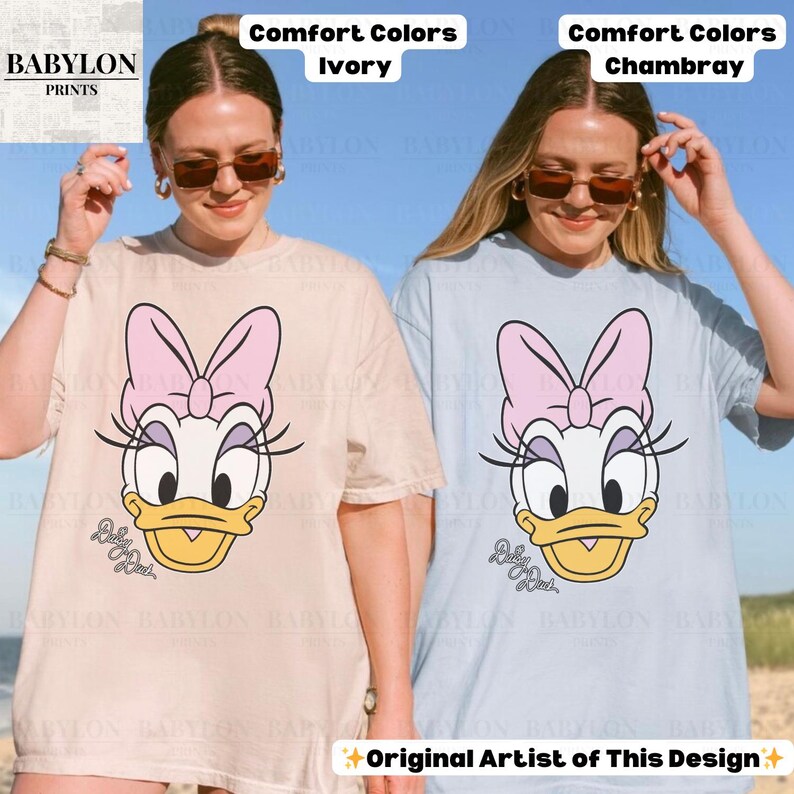 May include: Two t-shirts, one ivory and one chambray, each featuring a Daisy Duck graphic. The graphic includes a pink bow, eyelashes, and the text "Baby Duck". The shirts are labeled "Comfort Colors" and "Original Artist of This Design".