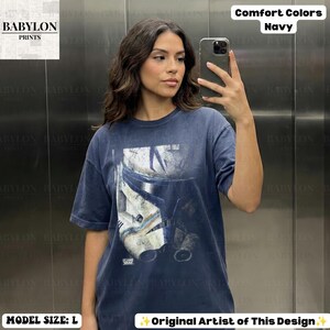 May include: A navy blue short-sleeve t-shirt featuring a distressed graphic print of a white and blue clone trooper helmet from Star Wars: The Clone Wars. The shirt has a crew neckline and a relaxed fit, with the text "CLONE WARS" printed below.