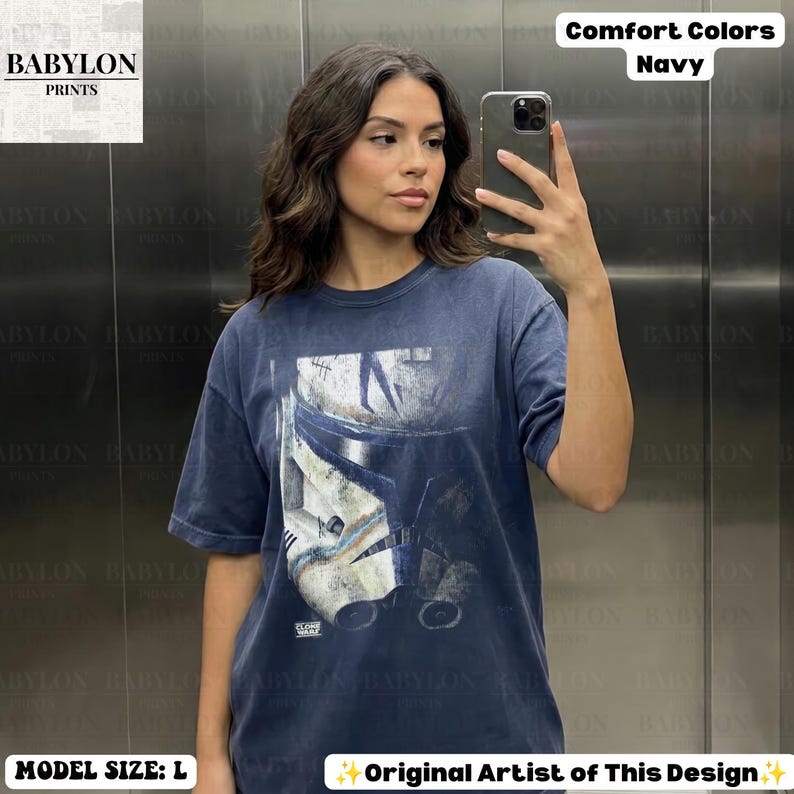 May include: A navy blue short-sleeve t-shirt featuring a distressed graphic print of a white and blue clone trooper helmet from Star Wars: The Clone Wars. The shirt has a crew neckline and a relaxed fit, with the text "CLONE WARS" printed below.