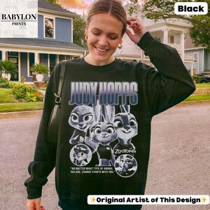 May include: Black sweatshirt featuring the text "JUDY HOPPS" and images of characters from the animated film "Zootopia." The design includes the film's title and the quote "NO MATTER WHAT TYPE OF ANIMAL YOU ARE, CHANGE STARTS WITH YOU."