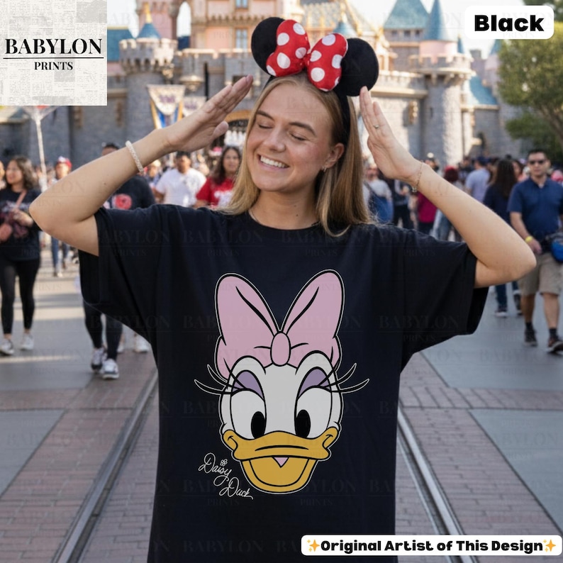 May include: Black t-shirt featuring a Daisy Duck graphic with a pink bow and yellow beak. The shirt has the text "Daisy Duck" in a cursive font. The model is wearing a Minnie Mouse ear headband with red polka dots.