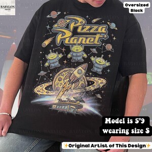 May include: Black oversized t-shirt featuring a colorful Pizza Planet graphic. The design includes the text "Pizza Planet", cartoon aliens, a rocket ship, and planets. The shirt is a size S.