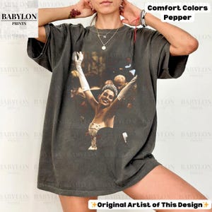 May include: Dark grey Comfort Colors t-shirt with a graphic of a woman wearing a tiara and gloves, arms raised. The shirt has the text "Original Artist of This Design" and "Comfort Colors Pepper".