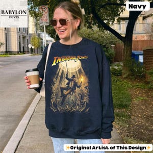 May include: Navy blue sweatshirt featuring the text "INDIANA JONES" in yellow and a graphic of the character. The design includes a spotlight effect and figures. The sweatshirt is a long-sleeved crew neck.