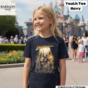 May include: Navy blue youth tee featuring the text "Indiana Jones" in yellow and the image of Indiana Jones. The graphic tee is worn by a young girl in front of a castle.