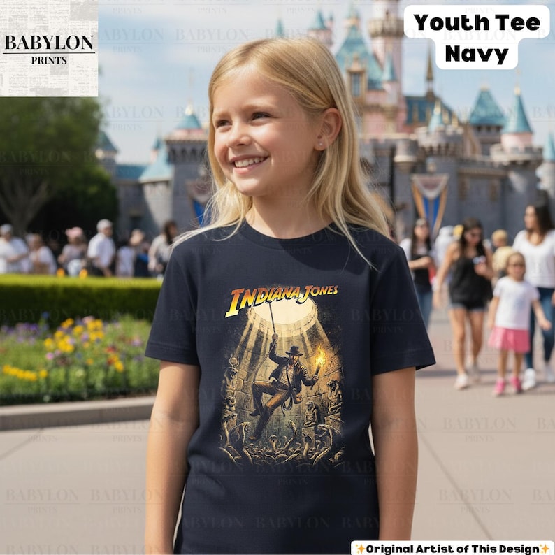 May include: Navy blue youth tee featuring the text "Indiana Jones" in yellow and the image of Indiana Jones. The graphic tee is worn by a young girl in front of a castle.