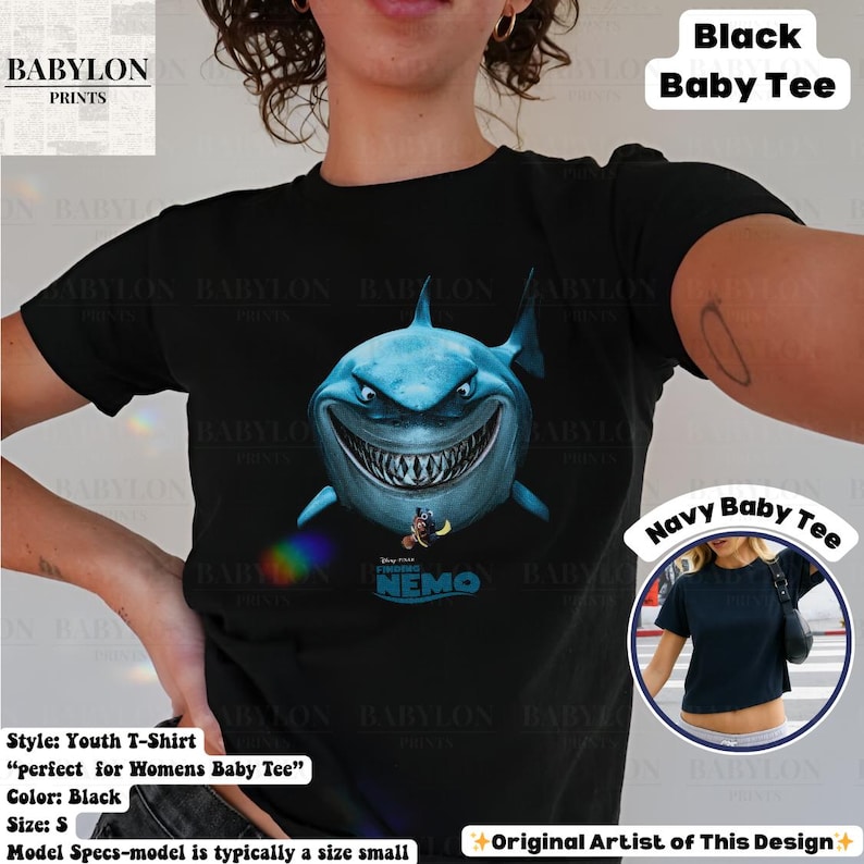 May include: Black youth t-shirt featuring a large, detailed illustration of a shark with sharp teeth. The shark is blue with a menacing grin, and the words "Finding Nemo" are below it. The shirt also has the text "Black Baby Tee".