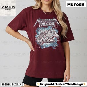 May include: Maroon t-shirt featuring a graphic of the Millennium Falcon with the text "Millennium Falcon". The shirt is a dark red color. The model is wearing a size XL. The design is by the original artist.
