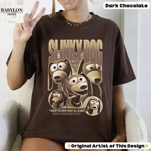 May include: Dark chocolate-colored t-shirt featuring a graphic of Slinky Dog from Toy Story. The design includes the text "SLINKY DOG" in a gold font, along with images of the character. The shirt also has the quote "I KNEW YOU WERE RIGHT ALL ALONG, WOODY! NEVER DOUBTED YA!"
