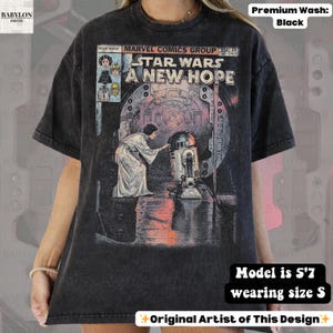 May include: Black t-shirt featuring a comic book-style graphic from Star Wars: A New Hope. The design includes Princess Leia and R2-D2. The shirt is a premium wash and the model is 5'7" wearing a size S.