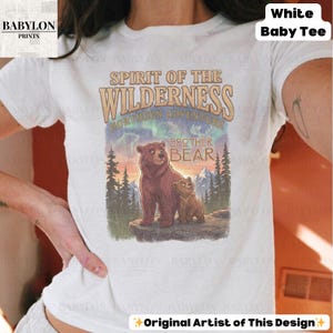 May include: White baby tee with a graphic of two bears and the text "Spirit of the Wilderness Northern Adventure Brother Bear." The design includes a mountain scene with trees and an aurora borealis effect. The shirt has the Babylon Prints logo.