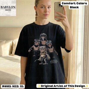Op de afbeelding: Een zwart t-shirt met korte mouwen en een grafische print van vier kleine, harige wezens met grote ogen en een bril. De tekst 'STAR WARS' staat in het wit onder de wezens gedrukt. Het shirt heeft een ronde hals en een relaxte pasvorm.