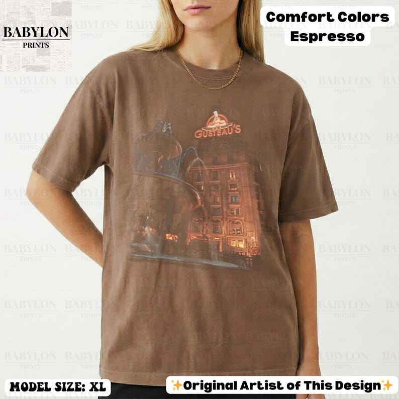 May include: Espresso-coloured t-shirt featuring a building graphic and the text "GUSTEAU'S". The shirt is from Babylon Prints and is a Comfort Colour. The model is wearing size XL. The design is by the original artist.