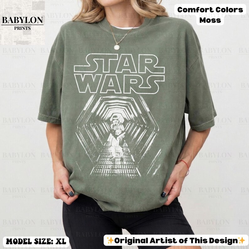 May include: Moss green Star Wars graphic t-shirt. The design features the Star Wars logo and a detailed illustration of a scene from the movie. The shirt is a comfort color and the model is wearing an XL size.