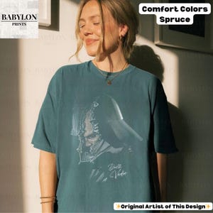 K&ouml;nnte beinhalten: Fichtefarbenes T-Shirt mit Darth Vader-Grafik. Das Design enth&auml;lt den Text "Darth Vader" in einer stilisierten Schriftart. Das Shirt geh&ouml;rt zur Comfort Colors-Kollektion und ist mit "Original Artist of This Design" gekennzeichnet.