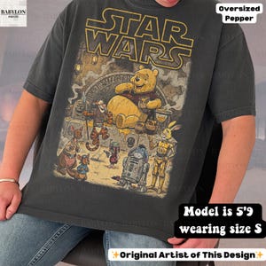 Może przedstawiać: Ciemnoszara, obszerna koszulka z grafiką Star Wars i Kubusia Puchatka. Projekt zawiera postacie takie jak Kubuś Puchatek, R2-D2 i inne. Tekst "STAR WARS" znajduje się na g&oacute;rze. Modelka ma 1,75 m wzrostu i nosi rozmiar S.