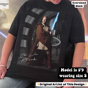 Może przedstawiać: Czarny, duży t-shirt z grafiką Obi-Wana Kenobiego trzymającego niebieski miecz świetlny. Tekst "Obi-Wan Kenobi" znajduje się poniżej. Koszulka w rozmiarze S, modelka ma 1,75 m wzrostu. Projekt oryginalnego artysty.