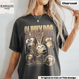 May include: Charcoal-colored t-shirt featuring a graphic of Slinky Dog from Toy Story. The design includes the text "SLINKY DOG" in gold lettering and several images of the character. The shirt also has the text "I KNEW YOU WERE RIGHT ALL ALONG, WOODY! NEVER DOUBTED YA!"