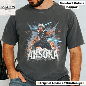 Może przedstawiać: Ciemnoszara koszulka z grafiką Ahsoki Tano z niebieskimi mieczami świetlnymi i napisem "AHSOKA". Koszulka wykonana jest z tkaniny Comfort Colors Pepper. Logo Babylon Prints znajduje się w lewym g&oacute;rnym rogu. Tekst "Original Artist of This Design" znajduje się na dole.