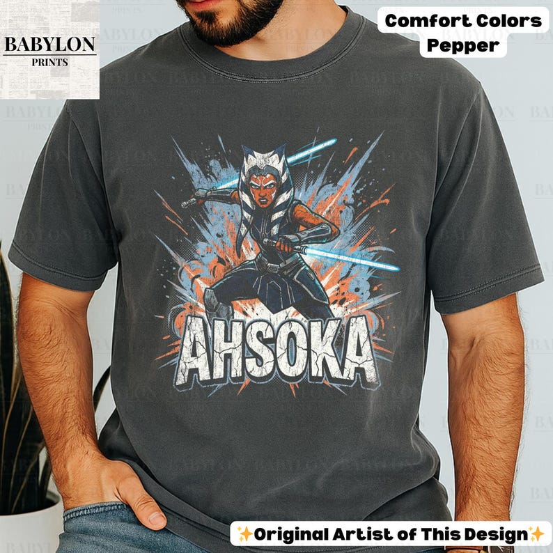 Może przedstawiać: Ciemnoszara koszulka z grafiką Ahsoki Tano z niebieskimi mieczami świetlnymi i napisem "AHSOKA". Koszulka wykonana jest z tkaniny Comfort Colors Pepper. Logo Babylon Prints znajduje się w lewym g&oacute;rnym rogu. Tekst "Original Artist of This Design" znajduje się na dole.