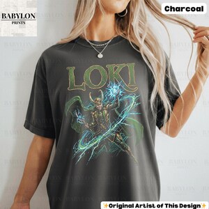 May include: Charcoal-colored t-shirt featuring the word "LOKI" in green and gold lettering. The shirt displays a graphic of Loki with green and blue lightning effects. The shirt is from Babylon Prints.