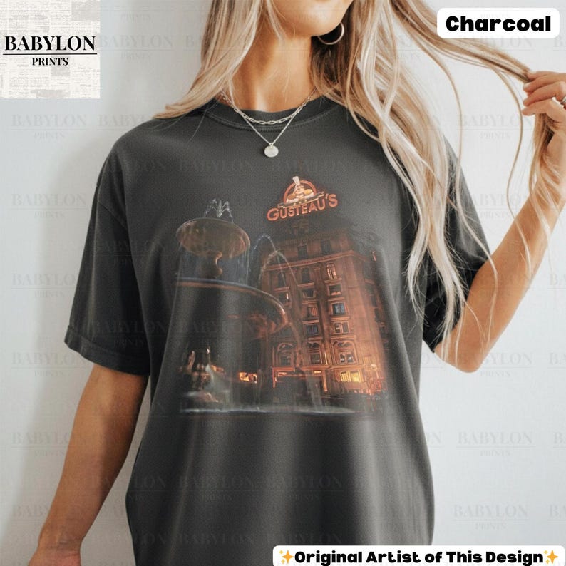 May include: A charcoal-coloured t-shirt with a graphic of a building and fountain, and the text "GUSTEAU'S". The shirt also has the text "Original Artist of This Design". The shirt is from Babylon Prints.