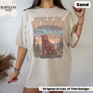 May include: Sand-coloured t-shirt with a graphic of two bears and the text "Spirit of the Wilderness" and "Brother Bear." The design includes a mountain landscape with trees and a colourful sky. The shirt has a relaxed fit.