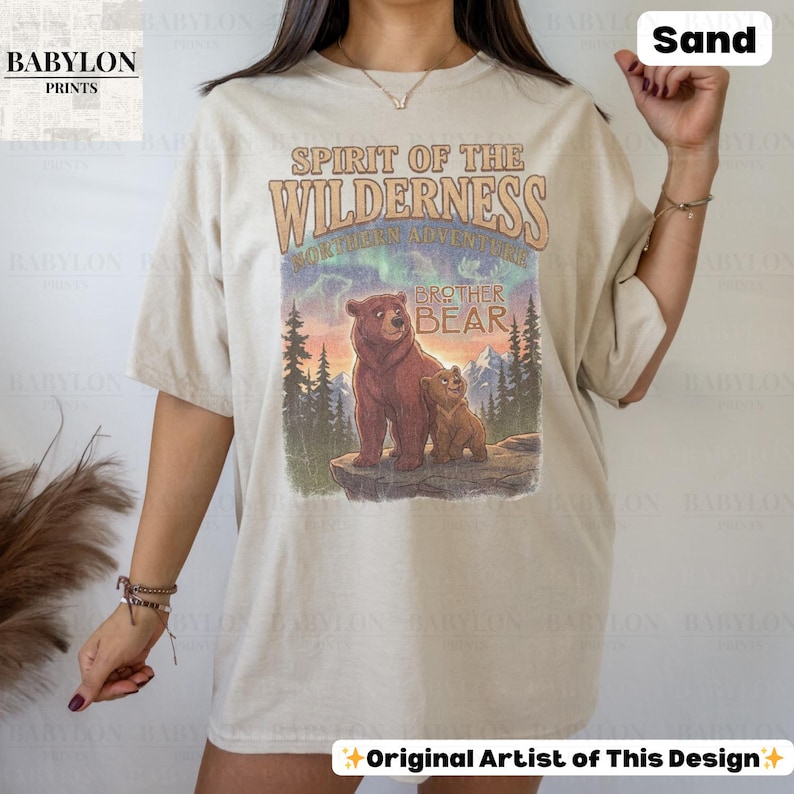 May include: Sand-coloured t-shirt with a graphic of two bears and the text "Spirit of the Wilderness" and "Brother Bear." The design includes a mountain landscape with trees and a colourful sky. The shirt has a relaxed fit.