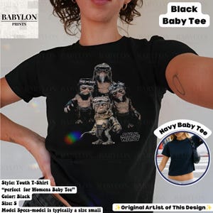 Op de afbeelding: Een zwart t-shirt met korte mouwen en een grafische print van vier kleine, harige wezens met een bril en een regenboogkleurig lichteffect. De tekst "STAR WARS" staat in het wit rechtsonder de afbeelding. Een kleine cirkelvormige inzet toont een marineblauwe versie van het t-shirt.