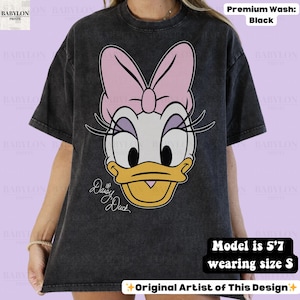 Puede incluir: Camiseta negra con un gr&aacute;fico de Daisy Duck, incluyendo un lazo rosa y un pico amarillo. El texto "Daisy Duck" est&aacute; escrito en cursiva. La camiseta es de lavado premium. La modelo mide 170 cm y usa talla S.