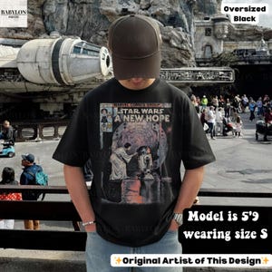 May include: Black oversized t-shirt featuring a comic book-style graphic from Star Wars: A New Hope. The shirt has short sleeves and is worn with a brown baseball cap. The text "STAR WARS A NEW HOPE" is visible on the front.