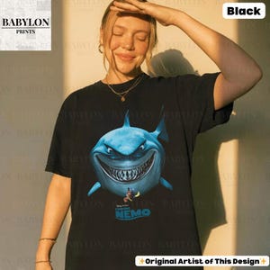 May include: Black t-shirt featuring a large, blue shark graphic from the movie "Finding Nemo." The shark has a menacing grin and is surrounded by the text "Disney Pixar Finding Nemo." The shirt also has the text "Original Artist of This Design."