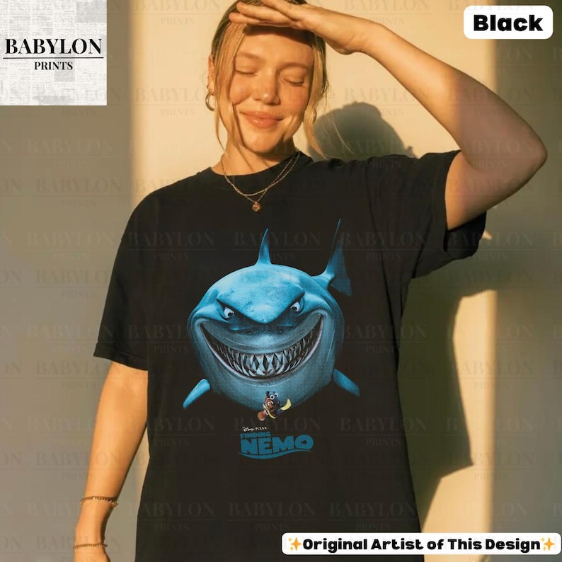 May include: Black t-shirt featuring a large, blue shark graphic from the movie "Finding Nemo." The shark has a menacing grin and is surrounded by the text "Disney Pixar Finding Nemo." The shirt also has the text "Original Artist of This Design."