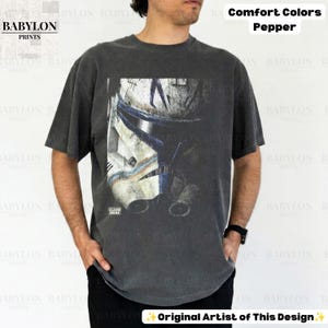 May include: A charcoal gray short-sleeve t-shirt featuring a large, distressed graphic print of a white and blue clone trooper helmet on the front. The shirt includes the text Comfort Colors Pepper and Babylon Prints in the image.