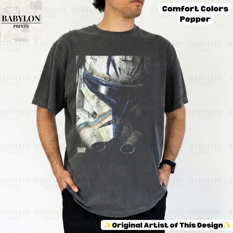 May include: A charcoal gray short-sleeve t-shirt featuring a large, distressed graphic print of a white and blue clone trooper helmet on the front. The shirt includes the text Comfort Colors Pepper and Babylon Prints in the image.