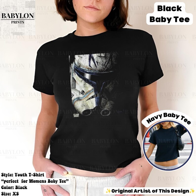 May include: A black short-sleeved baby tee featuring a graphic print of a weathered white and blue clone trooper helmet on the front. The shirt has a crew neckline and a cropped fit. A small circular inset shows a navy blue version of the same shirt.
