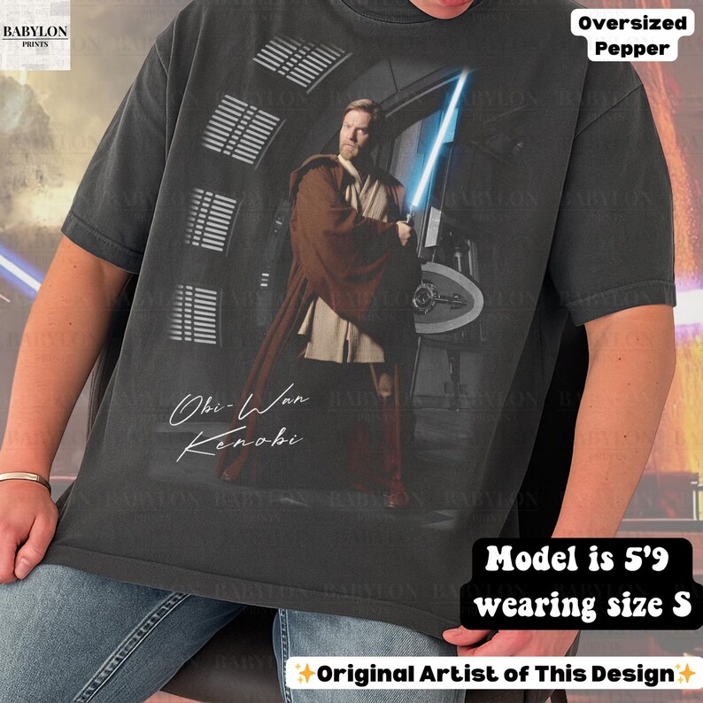 Może przedstawiać: Ciemnoszara koszulka oversize z grafiką Obi-Wana Kenobiego z mieczem świetlnym. Koszulka zawiera tekst "Obi-Wan Kenobi" i "Oversized Pepper". Modelka ma 1,75 m wzrostu i nosi rozmiar S. Original Artist of This Design.