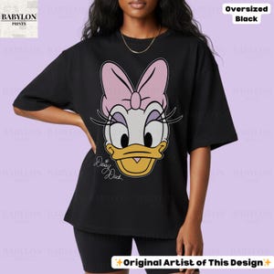 Puede incluir: Camiseta negra de gran tama&ntilde;o con la cara de dibujos animados de Daisy Duck y un lazo rosa. La camiseta tiene el texto "Daisy Duck" en una fuente de escritura. Las palabras "Oversized Black" est&aacute;n impresas en la parte superior derecha. El texto "Original Artist of This Design" est&aacute; impreso en la parte inferior.