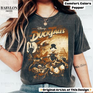 May include: A dark gray t-shirt featuring a vintage-style graphic from Disney's DuckTales. The design includes various characters and the show's logo in a distressed, sepia-toned print. The shirt has the text "Comfort Colors Pepper" and "Original Artist of This Design".